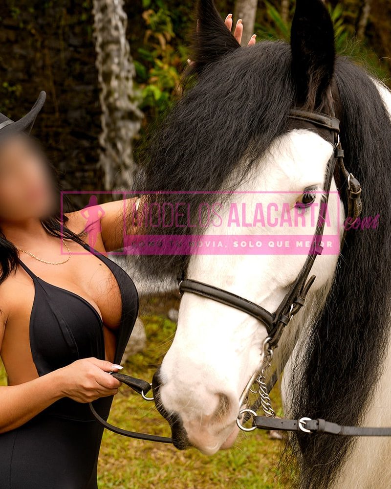 Mujer madura rica en Medellín, Naty Gold, junto a un caballo en un campo abierto, destacando la elegancia natural y la libertad en un entorno tranquilo.